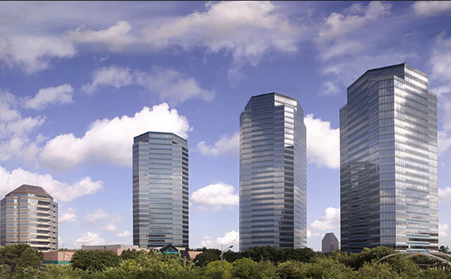 Glass skyscrapers form a modern city skyline against a blue sky with scattered clouds, with greenery in the foreground.
